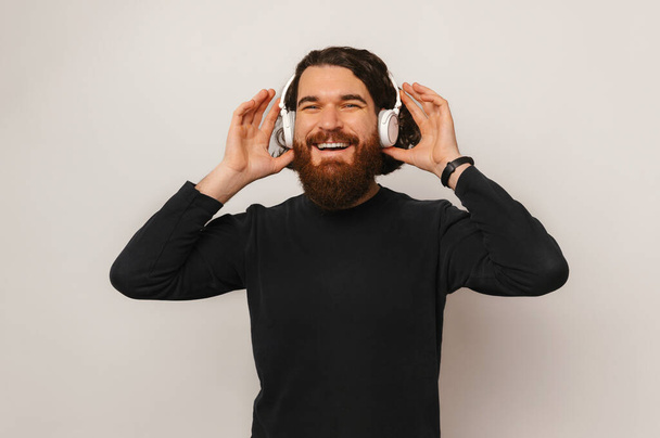 Joyful young bearded man with long hair listening music at wireless headphones over white background. - Foto, immagini