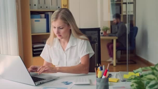 Mujer concentrada haciendo informe de negocios en el ordenador portátil, mientras que su compañero de trabajo sentado en segundo plano en una amplia oficina moderna - Imágenes, Vídeo
