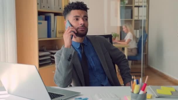Середній знімок молодого офісного менеджера, який розмовляє на мобільному телефоні з клієнтом під час угоди за столом, жінка-співробітниця на задньому плані - Кадри, відео