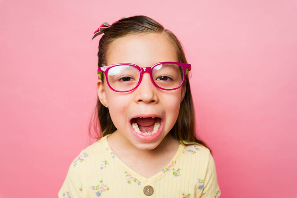 Nahaufnahme eines entzückenden kaukasischen kleinen Mädchens mit verschreibungspflichtiger Brille, das ihr großes zahnloses Lächeln zeigt, das niedlich schreiend aussieht - Foto, Bild