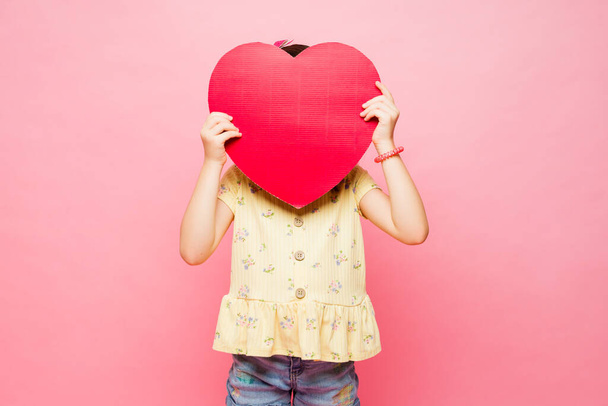 Любящая старшеклассница прячет свое лицо за большим красным сердцем и дает любовь семье и друзьям на розовом фоне - Фото, изображение