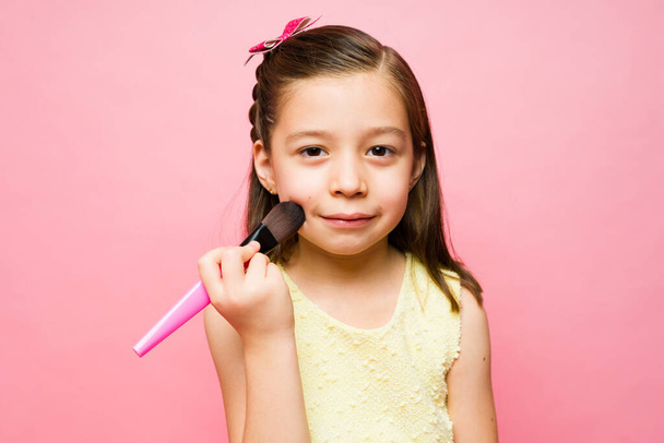 Portrét roztomilé rozkošné základní věk holčička hraje s make-up pomocí štětce při vytváření očního kontaktu v růžovém pozadí - Fotografie, Obrázek