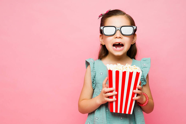 Kaukasisches kleines Kind sieht überrascht und aufgeregt aus, als es sein zahnloses Lächeln mit einer 3D-Brille zeigt und einen Film mit Popcorn anschaut - Foto, Bild