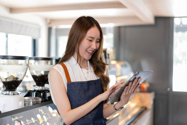 Young Asian female coffee shop owner and barista using a digital tablet. Millennial Asia woman smiling and working at cafe and restaurant indoors. Technology usage  - Foto, Imagen