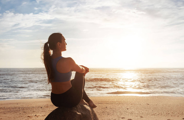 Spokojny pozytywny młoda kobieta kaukaska siedzieć na kamieniu, cieszyć się porannym treningiem, letni wschód słońca, odpoczynek, relaks od treningu na plaży morskiej. Przerwa od sportu, stylu życia i opieki zdrowotnej na świeżym powietrzu - Zdjęcie, obraz