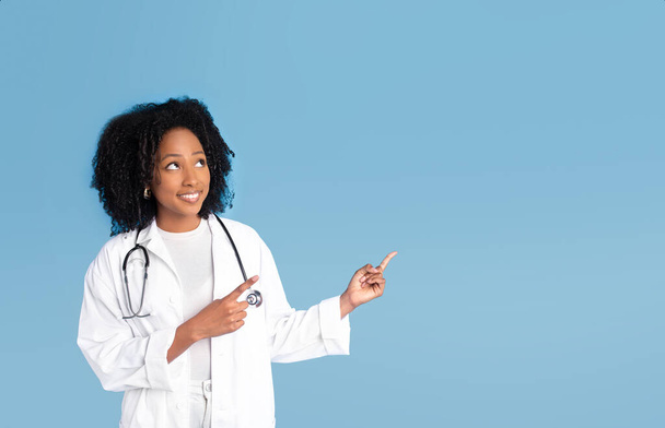 Boldog szép fekete millenniumi hölgy orvos fehér kabátban ujjal felfelé szabad hely, azt javasolja, hirdetés és ajánlat, elszigetelt kék stúdió háttér. Szakmai tanácsadás, értékesítés, orvostudomány, egészségügy - Fotó, kép
