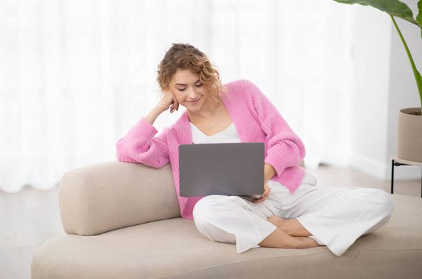 Domowy wypoczynek. Uśmiechnięta młoda dama z laptopem relaksująca się na kanapie w domu, piękna Tysiąclecia kobieta korzystająca z komputera w salonie, oglądająca filmy lub przeglądająca Internet, ciesząca się weekendową rozrywką - Zdjęcie, obraz
