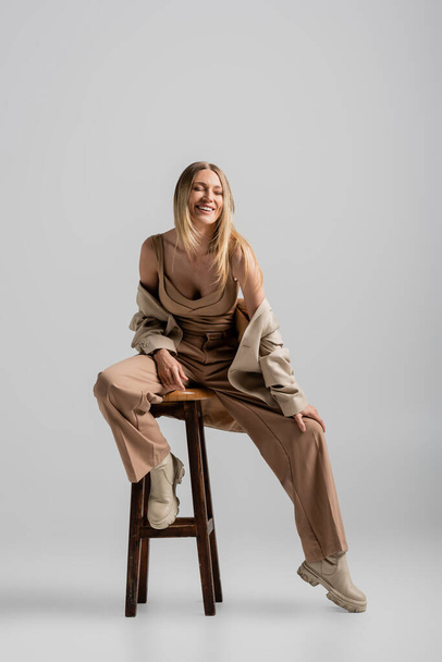 vrolijke blonde vrouw in stijlvolle formele pak zitten op stoel en glimlachen, mode en stijl - Foto, afbeelding