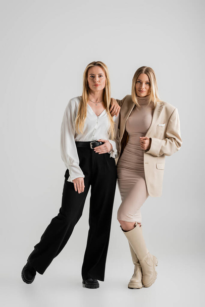 stijlvolle blonde zussen in formele outfit samen poseren op grijze achtergrond, fashion concept - Foto, afbeelding