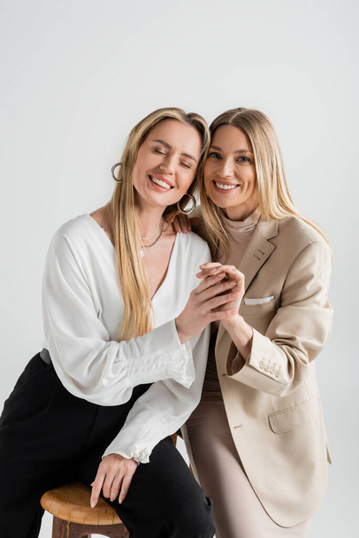 two attractive elegant sisters sitting on chairs smiling and holding hands, fashion and style - Fotografie, Obrázek