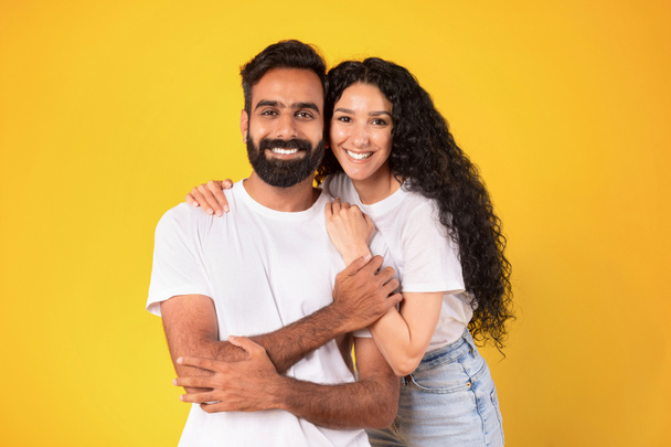 Romantic Couple in love. Portrait of loving young arabic spouses hugging standing together over yellow studio background. Happy woman and man sharing embrace enjoying their relationship - Photo, Image