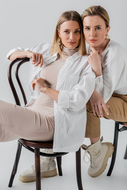 stylish elegant blonde sisters sitting on chairs in fashionable pastel attire, fashion concept - Photo, Image