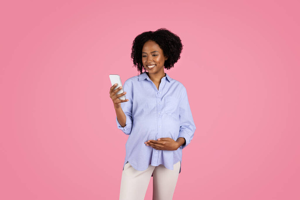 Cheerful young black pregnant woman in casual touch belly, use smartphone, chatting in social networks, isolated on pink studio background. App, device for expect baby, ad and offer - Photo, Image