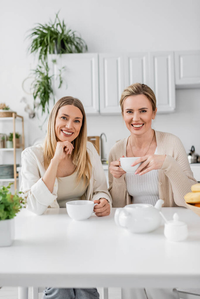 zwei Schwestern in pastellfarbenem Outfit, die lächelnd Tee trinken und in die Kamera schauen, Familienbande - Foto, Bild