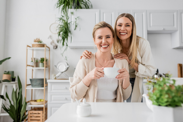 zwei blonde Schwestern trinken Tee und lächeln auf Küchenhintergrund mit Pflanzen, familiäre Bindung - Foto, Bild