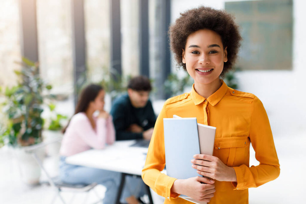 Портрет щасливої чорної леді студентки університету позує з блокнотами і посміхається, коли її однокласники навчаються на фоні сучасного інтер'єру аудиторії - Фото, зображення