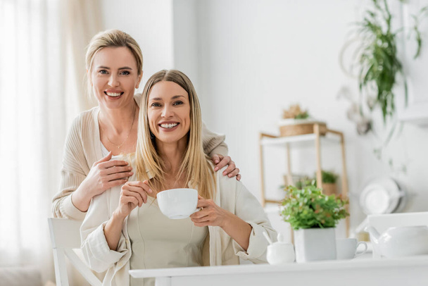 twee vrolijk mooi zussen kijken naar camera glimlachen en poseren op keuken achtergrond, familie binding - Foto, afbeelding