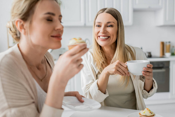 close up two beautiful sisters in casual wear tasting cupcakes and drinking tea, family bonding - Photo, Image
