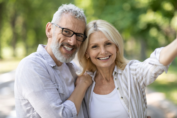Happy beautiful mature couple posing for selfie together outdoors, cheerful senior man and woman talking photo while walking in park, smiling and looking at camera, enjoying date outside, closeup - Photo, Image