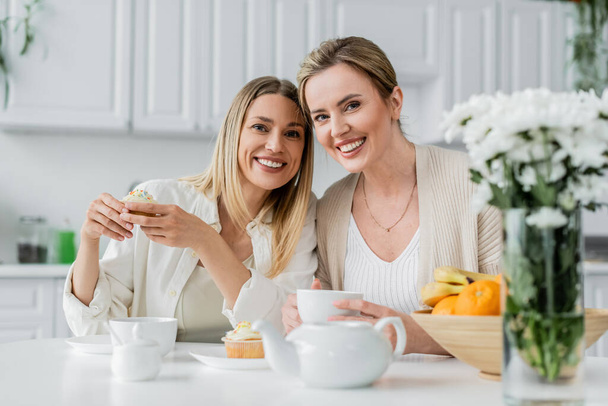 alegre encantadora hermanas en traje de moda disfrutando de té y cupcakes y mirando a la cámara, vinculación - Foto, Imagen