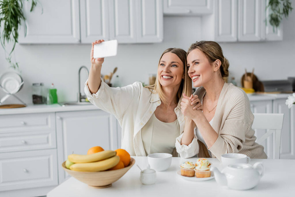 classy sisters in trendy pastel cardigans making selfie at table in kitchen, family bonding - Photo, Image