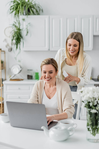 two blonde sisters in trendy attire looking at laptop on kitchen background, family bonding - Foto, afbeelding