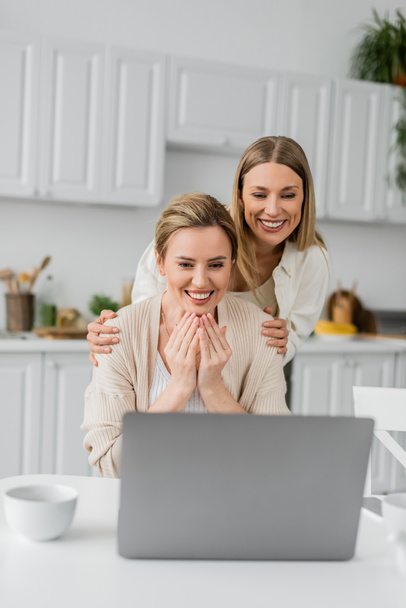 улыбающиеся сестры в красивых случайных кардиганах глядя на ноутбук, качество времени и семейной связи - Фото, изображение