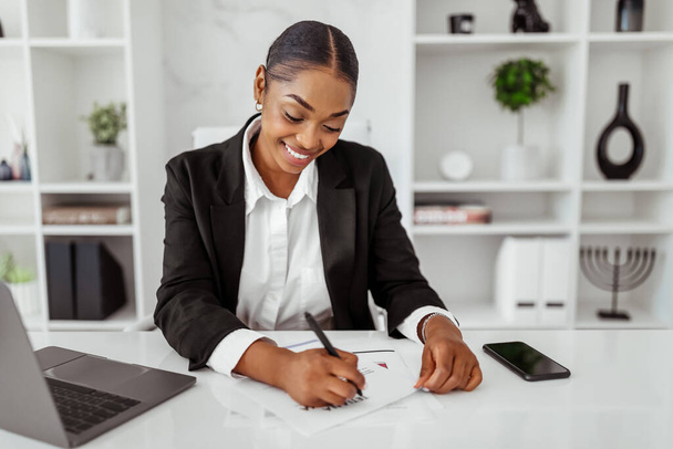 Boldog fekete női üzleti elemző, aki papírokra ír, adatokat, táblázatokat és grafikonokat ellenőriz, világos irodában ül az asztalnál. Női monitoring vállalati növekedés, nyereség és elemzés - Fotó, kép