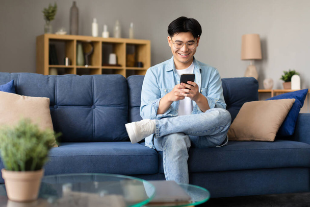 Happy asian guy in eyeglasses sitting with smartphone in hands, websurfing internet and browsing social networks on mobile phone, using application and messaging at home. Gadgets conceito de lazer - Foto, Imagem