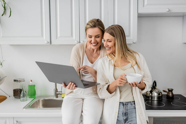 attractive jolly sisters in lovely outfit looking at laptop on kitchen furniture backdrop, bonding - Photo, Image