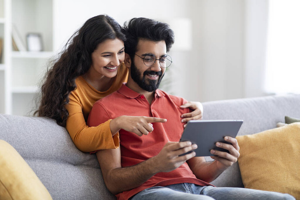 Tolle App. Junges indisches Paar nutzt digitales Tablet im heimischen Wohnzimmer, lächelnde Ost-Ehepartner surfen auf modernem Gerät im Internet, machen gemeinsam Online-Einkäufe, Frau zeigt auf Bildschirm - Foto, Bild