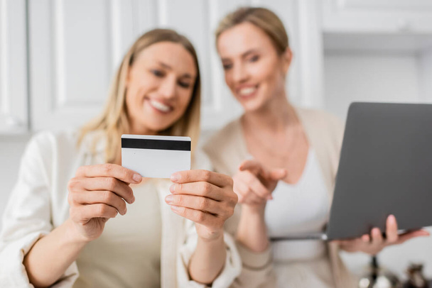 primer plano bien parecido hermanas de moda en la cocina con el ordenador portátil que sostiene la tarjeta de crédito, borrosa, vinculación - Foto, imagen