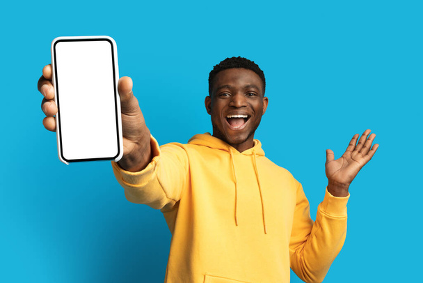 Izgalmas jóképű fiatal fekete srác sárga kapucnis mutatja nagy telefon fehér üres képernyő és gesztikuláló, ajánló szép online ajánlat, mobil app, elszigetelt kék háttér, másolás hely, mockup - Fotó, kép
