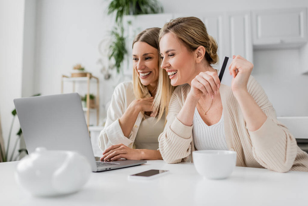 deux sœurs blondes gaies tenant la carte de crédit regardant l'ordinateur portable et souriant sincèrement, collant - Photo, image