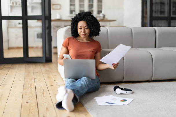 African American Freelancer Woman Working With Laptop And Documents At Home, Smiling Young Black Female Sitting On Floor In Room, Reading Papers And Using Computer, Enjoying Remote Work, Copy Space - Photo, Image