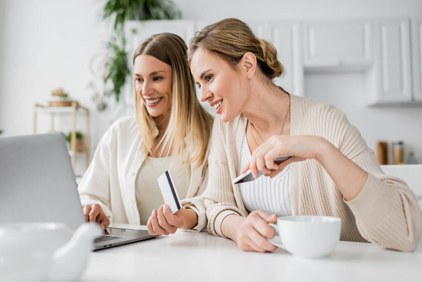 two lovely blonde sisters in trendy attire with credit card and tea cup looking at laptop, bonding - Photo, Image