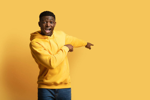 Excited handsome young african american guy in casual outfit pointing at copy space for advertisement behind him and grimacing, isolated on yellow background. Great offer, amazing deal - Foto, Imagem
