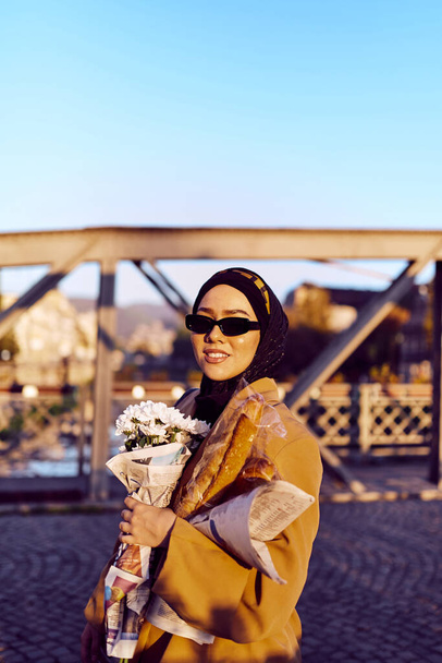 Eine Hijab-Frau mit stylischer Sonnenbrille und elegantem französischen Outfit spaziert bei Sonnenuntergang durch die Stadt, trägt einen Strauß, Brot und Zeitungen bei sich und strahlt einen Sinn für kulturellen Charme und Gelassenheit aus..  - Foto, Bild