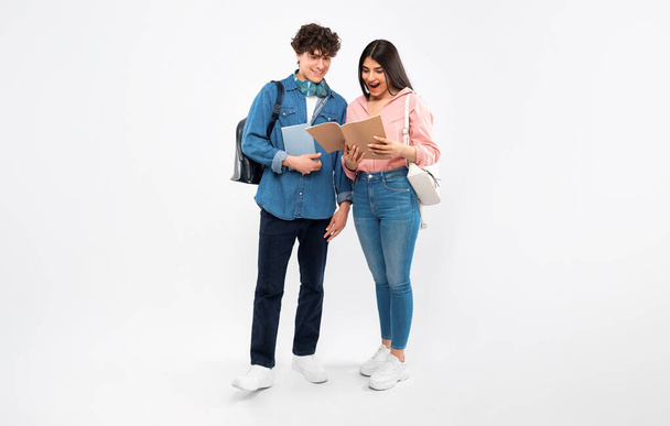 Academic Excellence. Joyful Students Couple Celebrating Academic Success Looking At Textbooks With Joy, Receiving Excellent Grades At College, Posing With Books And Backpacks On White Background - 写真・画像