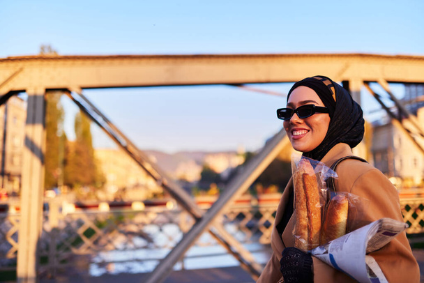 Una mujer hijab con gafas de sol elegantes y un elegante atuendo francés, caminando por la ciudad al atardecer, llevando un ramo, pan y periódico, irradiando un sentido de encanto cultural y serenidad..  - Foto, imagen