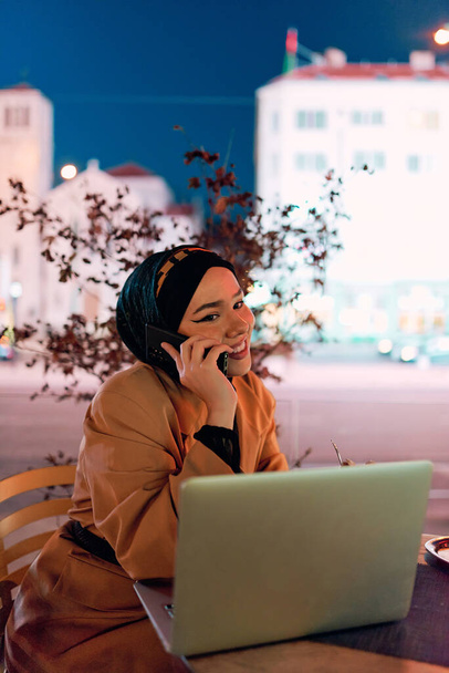Uma mulher hijab usando laptop e smartphone em um café moderno na paisagem urbana noturna, refletindo a mistura harmoniosa de tecnologia e ambiente urbano.  - Foto, Imagem