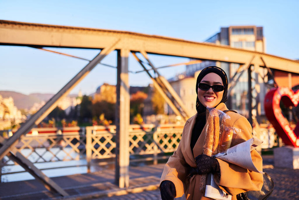 Женщина в хиджабе в стильных солнцезащитных очках и элегантном французском наряде, прогуливаясь по городу на закате солнца, неся букет, хлеб и газету, излучая чувство культурного шарма и спокойствия.  - Фото, изображение