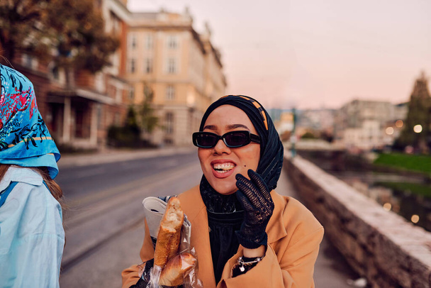 Пара женщин одета в хиджаб и современное, но традиционное платье, а другая в синем платье и шарфе, прогуливаясь вместе по городу на закате. Один несет букет и хлеб, в то время как - Фото, изображение