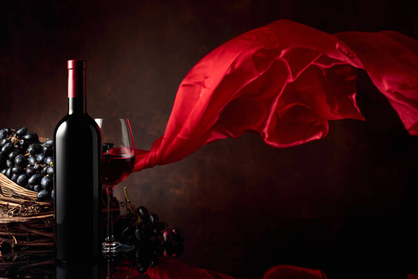 Glas en fles rode wijn met druiven op een zwarte reflecterende achtergrond. Rood satijnen gordijn wappert in de wind. Kopieerruimte. - Foto, afbeelding