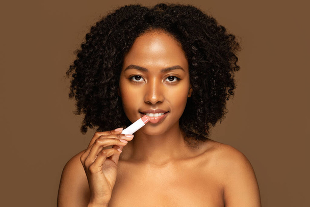 Portrait de beauté de souriante jolie jeune femme noire seins nus avec une peau parfaite et maquillage naturel sur fond brun tenant rouge à lèvres nu ou baume à lèvres. Soin des lèvres, concept maquillage - Photo, image