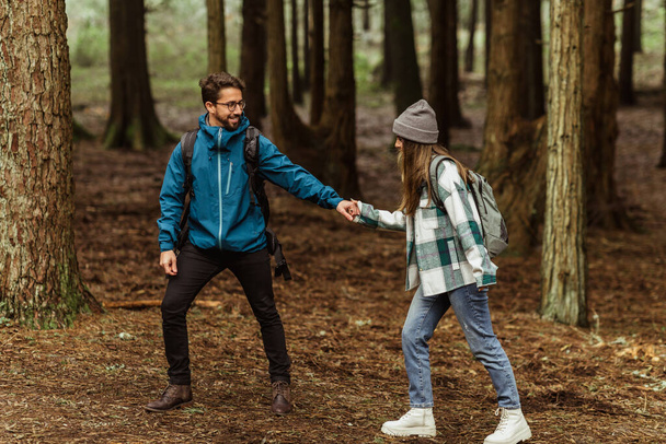 Cheerful young caucasian couple in jackets in forest together, guy helps to lady, hold hand, enjoy trip, outdoor. Hiking, adventure and tourism, active lifestyle, full length - Photo, Image