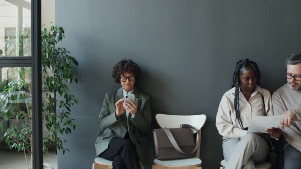 Μέση πλήρης βολή της καυκάσιας γυναίκας χρησιμοποιώντας smartphone, ενώ η μαύρη γυναίκα και ο Καυκάσιος άνδρας συζητούν έγγραφο, όλοι περιμένουν για συνέντευξη - Πλάνα, βίντεο