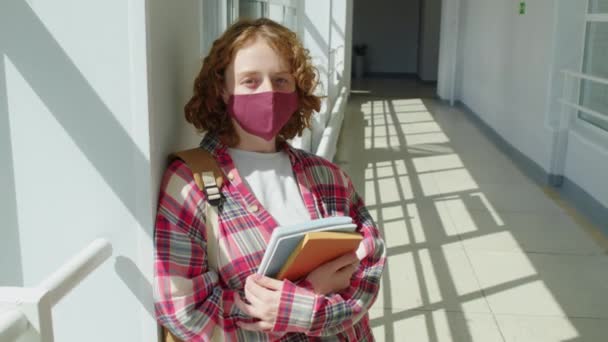 Keskikokoinen muotokuva tunnistamaton punatukkainen tyttö yllään suojaava naamio tilalla oppikirja ja copybooks seisoo koulun käytävällä katsomassa kameraa - Materiaali, video
