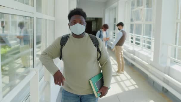 Portrait moyen d'un étudiant afro-américain portant un masque de protection tenant un manuel et un copybook debout dans un couloir universitaire regardant une caméra - Séquence, vidéo