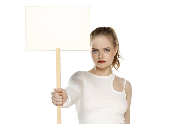 Mladé blondýny naštvaný žena drží prázdný znak na bílém pozadí studia. - Fotografie, Obrázek